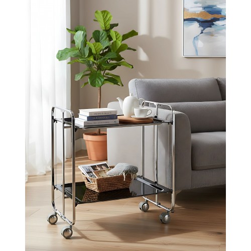 2-Tier FoldingKitchen Cart on Wheels – Chromed Frame and black shelves holding coffee cup, coffee maker and books next to a sofa